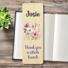 A personalized wooden bookmark with a light beige wood-grain finish, a watercolour floral design in the centre with purple, pink, and white flowers, and the text 'Thank you a whole bunch' in purple at the bottom. The bookmark is displayed on a rustic wooden tabletop next to an open notebook and a small green succulent.