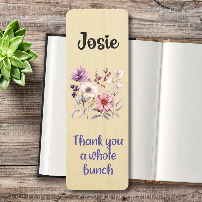 A personalized wooden bookmark with a light beige wood-grain finish, a watercolour floral design in the centre with purple, pink, and white flowers, and the text 'Thank you a whole bunch' in purple at the bottom. The bookmark is displayed on a rustic wooden tabletop next to an open notebook and a small green succulent.