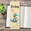 A personalized wooden bookmark with a light beige wood-grain finish, featuring a cute blue-green cartoon dinosaur, a small orange bird, and the name 'Erika' printed in black. The bookmark is displayed on a rustic wooden tabletop next to an open notebook and a green succulent.