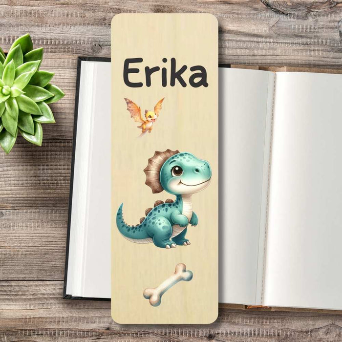 A personalized wooden bookmark with a light beige wood-grain finish, featuring a cute blue-green cartoon dinosaur, a small orange bird, and the name 'Erika' printed in black. The bookmark is displayed on a rustic wooden tabletop next to an open notebook and a green succulent.