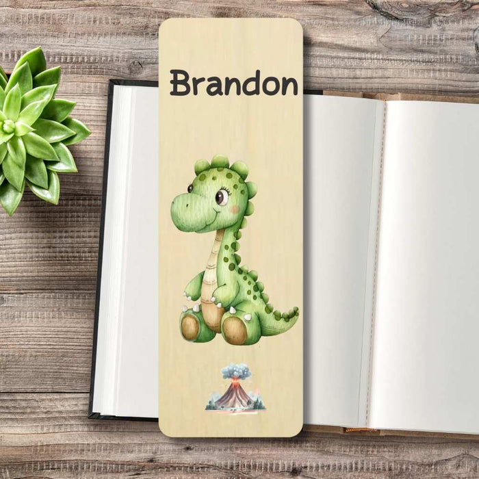 A personalized wooden bookmark featuring a cute green cartoon dinosaur displayed on an open notebook over a rustic wood background with a small succulent plant.
