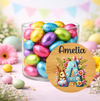 A clear glass jar filled with colorful foil-wrapped Easter eggs, styled on a pastel pink surface with decorative flowers, pastel eggs, and a round custom wooden name plaque reading 'Amelia' with a floral letter A and bunny illustration.