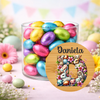 A clear glass jar filled with colorful foil Easter eggs, surrounded by pastel flowers and decorative Easter props. In front of the jar is a round wooden-style personalized label with the name 'Daniela' above a large floral bunny letter 'D'.