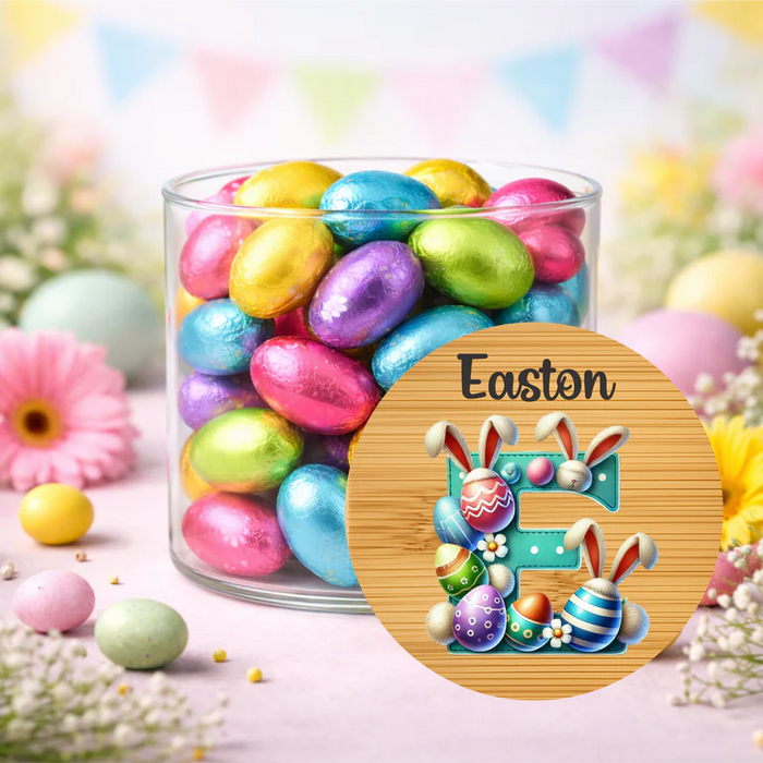 A clear glass jar filled with pastel foil Easter eggs, surrounded by decorative flowers, pastel eggs, and bunting. A round wooden-style overlay in front shows the text 'Easton' above an illustrated Easter egg and bunny design.