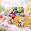 Jar of colorful Easter eggs with a bunny and egg design on a wooden circle, surrounded by flowers.