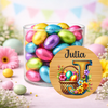 A clear glass jar filled with colorful foil-wrapped chocolate eggs, set on a pastel pink surface with decorative flowers and bunting in the background. A large round personalized monogram sticker reading 'Julia' is overlaid in the foreground.
