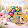 A clear glass jar filled with colorful foil-wrapped chocolate eggs on a pastel Easter-themed background, with a round personalized label in the foreground reading 'Leah' and featuring a large number 1 with a bunny illustration.