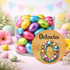 A clear glass jar filled with colorful foil Easter eggs, set on a pastel pink surface with decorative flowers and bunting in the background. A round wooden-style label in front reads 'Octavia' above a large floral number 0.