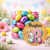 A clear glass jar filled with colorful foil-wrapped Easter eggs, set on a pastel pink surface with flowers and bunting in the background. A round wooden-style label in front reads 'Payton' above a large pink letter P with a white bunny and a pink polka-dotted egg.