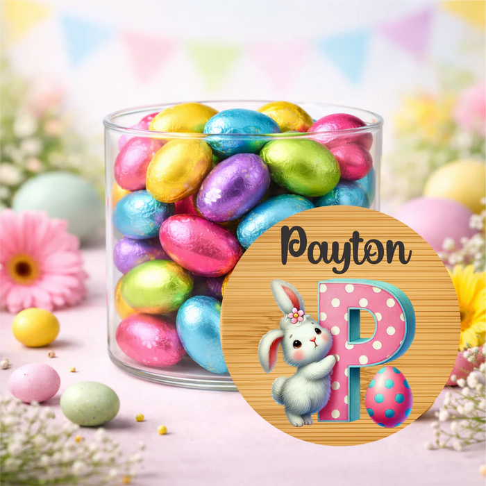A clear glass jar filled with colorful foil-wrapped Easter eggs, set on a pastel pink surface with flowers and bunting in the background. A round wooden-style label in front reads 'Payton' above a large pink letter P with a white bunny and a pink polka-dotted egg.