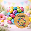A clear cylindrical glass jar filled with colorful foil-wrapped chocolate eggs, set on a pastel surface with decorative flowers and Easter-themed props. A round wooden-style label in front reads 'Quincy' above a floral wreath design.
