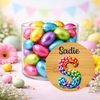A clear cylindrical glass jar filled with colorful foil-wrapped chocolate eggs, set on a pastel surface with flowers and bunting in the background. A round wooden-style label in front reads “Sadie” above a large rainbow-colored decorative letter S.