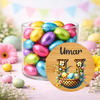 A clear glass jar filled with colorful foil-wrapped chocolate eggs, styled with pastel flowers and Easter decorations. A round wooden-style label overlay in front reads 'Umar' with a decorative letter H and basket of eggs.