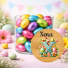 A clear glass jar filled with colorful foil-wrapped Easter eggs, surrounded by pastel flowers and eggs on a pink surface. A round wooden-style label overlay in front reads 'Xena' above a large decorative letter X with floral and bunny details.