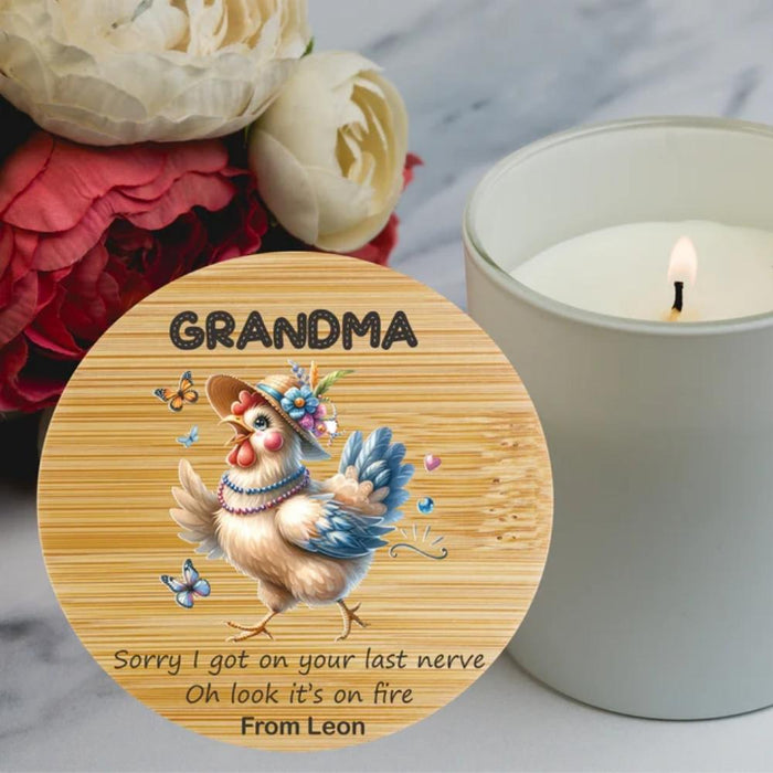 A personalized round wooden plaque or coaster featuring an illustration of a hen wearing a hat and flower crown, with the text 'GRANDMA' at the top and a humorous quote at the bottom. The plaque is styled on a marble surface next to a lit white candle and flowers.