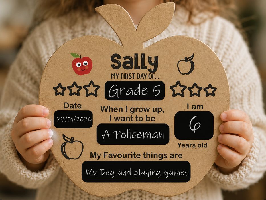 A child in a cream knit sweater holds a large apple-shaped cardboard sign with personalized school and life milestones printed on it, including the name, Grade 5, date, age, and a favorite phrase.