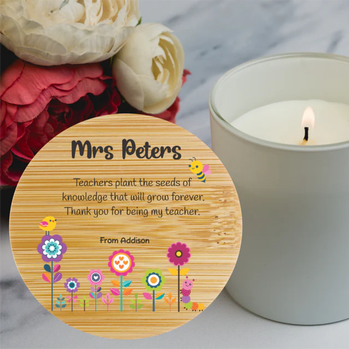 A round wooden keepsake plaque with a personalized message for a teacher, displayed next to a lit white candle and flowers on a marble surface.