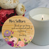 A round wooden candlestick coaster or label with floral artwork and the text saying thank you to a teacher is shown in front of a lit white candle on a light marble surface, with flowers in the background.
