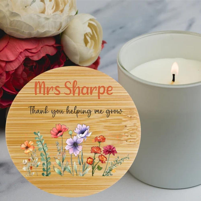 A round wooden bamboo lid for a candle with the personalized text 'Teacher Thank you helping me grow' and a colourful wildflower illustration, styled next to a lit white candle and flowers on a marble surface.