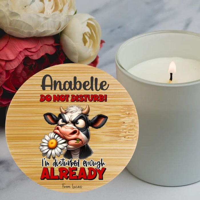 A round wooden plaque or coaster with a cartoon angry cow holding a daisy, surrounded by the personalised text ' DO NOT DISTURB! I'm disturbed enough ALREADY' in black and red lettering. The plaque is styled on a white marble surface next to a lit white candle and red and white flowers.