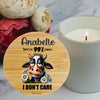 A round wooden bamboo lid for a candle with a personalized design featuring a cartoon cow and the personalised text 'friend there's a 99% chance I DON'T CARE' is shown in the foreground next to a lit white candle on a marble surface with flowers in the background.