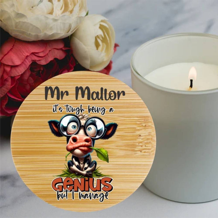 A round wooden bamboo lid for a candle featuring a funny cartoon of a cow wearing glasses and holding a leaf, with the personalised text for a teacher ' it's tough being a Genius but I manage'.