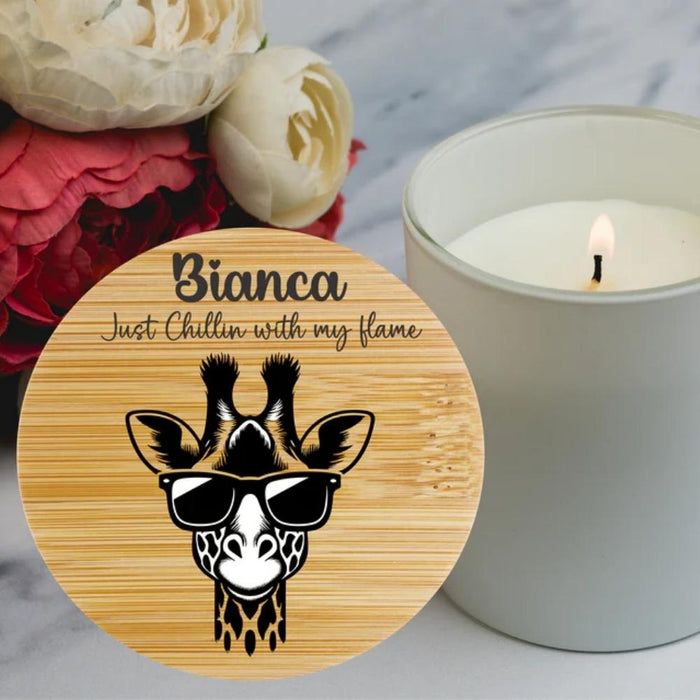 A round wooden bamboo lid for a candle featuring a cartoon giraffe wearing black sunglasses and the personalised text for a sister 'Just Chillin with my flame', displayed next to a lit white candle in a glass jar on a marble surface with flowers in the background.