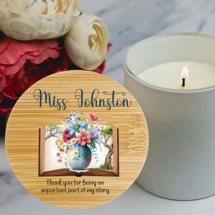A personalized wooden keepsake bamboo lid for a candle, featuring personalised name in script and an illustration of an open book with colorful flowers and a blue vase. The item is styled on a marble surface next to a lit white candle and red and cream flowers in the background.