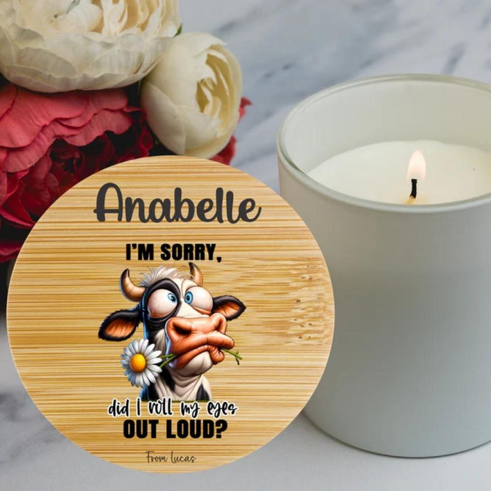 A round bamboo lid with a custom printed design featuring a cartoon cow and the personalised text ' I'M SORRY, did I roll my eyes OUT LOUD?'. It is shown next to a lit white candle on a marble surface with flowers in the background.