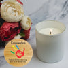 A lit white candle in a frosted glass jar sits on a light marble surface beside cream and red flowers. A round bamboo-style label in the lower left shows personalised text and colourful handprint artwork with a thank-you message.