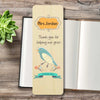 A personalized wooden bookmark with a bird illustration and the text 'Mrs Jordan Thank-you for helping me grow' and 'THANK YOU', displayed on a rustic wooden table next to an open book and a small succulent.