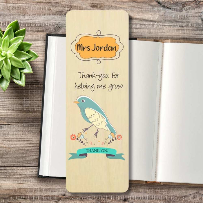 A personalized wooden bookmark with a bird illustration and the text 'Mrs Jordan Thank-you for helping me grow' and 'THANK YOU', displayed on a rustic wooden table next to an open book and a small succulent.