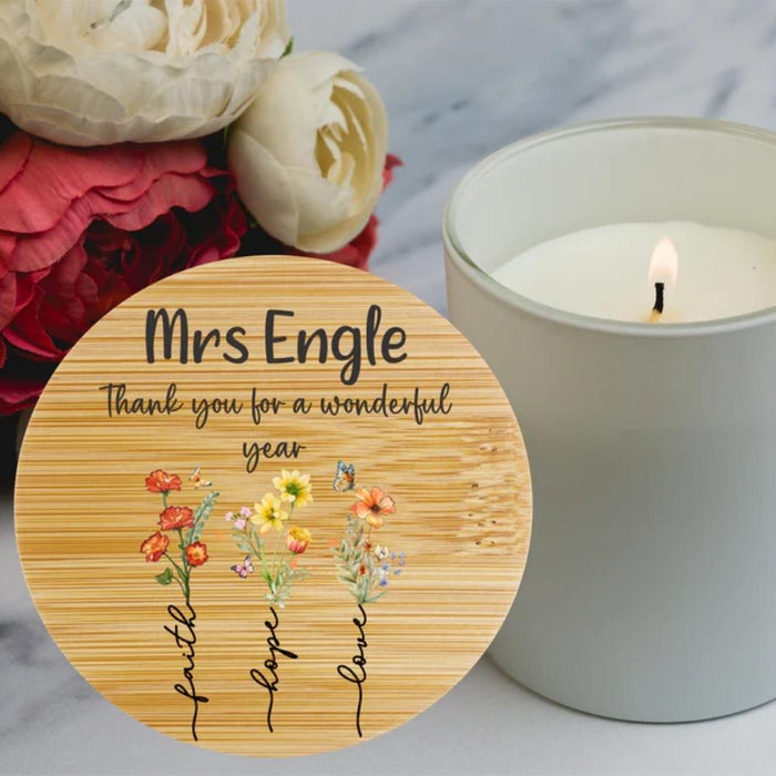 A round wooden bamboo lid candle engraved with 'Thank you for a wonderful year' and small floral illustrations, styled beside a lit white candle in a glass jar with flowers in the background.