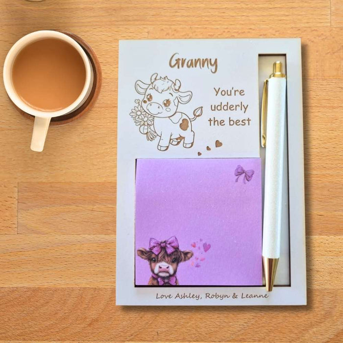 A personalized stationery gift set on a wooden table, featuring a white card with a cartoon cow illustration, the text 'You're udderly the best', a lavender sticky note with a cow and bow design, and a white pen with gold accents. A cup of coffee is placed to the left.