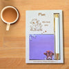 A personalized Mother's Day gift set on a wooden table, featuring a white card with a cow illustration and 'We love you' message, a purple sticky note with a cow print, and a white pen in a slot.