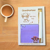 A personalized stationery gift set on a wooden table, featuring a cream-colored notebook with a cow illustration and the text 'Grandmamaaa We love You', a purple notepad with a cow illustration, and a white pen with gold accents.