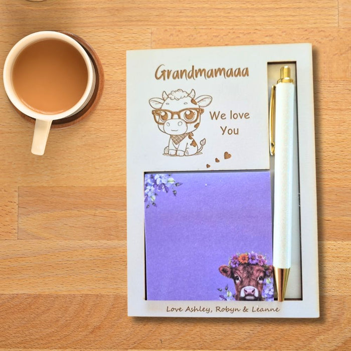 A personalized stationery gift set on a wooden table, featuring a cream-colored notebook with a cow illustration and the text 'Grandmamaaa We love You', a purple notepad with a cow illustration, and a white pen with gold accents.