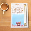 A personalized mother's day gift set on a light wood table, featuring a white card with cute cow illustrations and the text 'Mom Have you heard, we have the best mom', a blue floral notepad, and a white pen with gold accents in a slot. A cup of coffee is visible in the top left corner.