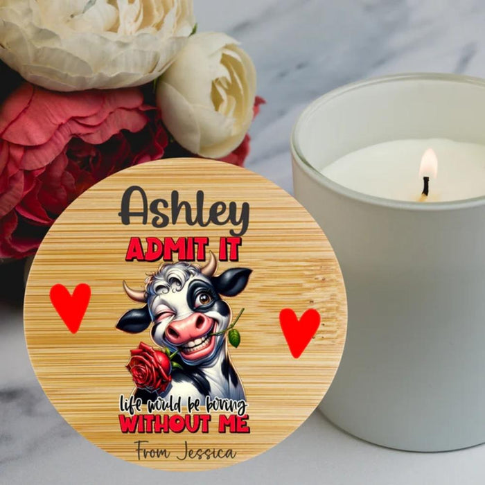 A round wooden bamboo lid for a candle in a glass jar with a custom funny cow illustration and the text 'ADMIT IT' and 'life would be boring WITHOUT ME' is shown on a marble surface next to a lit candle and flowers.