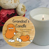 A personalised round  wooden candle tin labelled with a cartoon orange character and the text 'To be lit in case of emergency' and 'Love from The Gang', shown beside a lit white candle on a marble surface with flowers in the background.