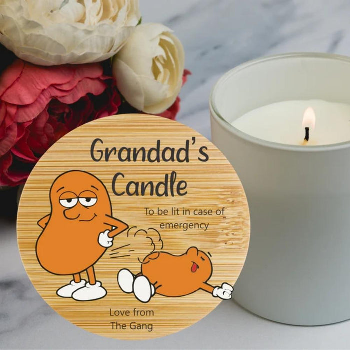 A personalised round  wooden candle tin labelled with a cartoon orange character and the text 'To be lit in case of emergency' and 'Love from The Gang', shown beside a lit white candle on a marble surface with flowers in the background.