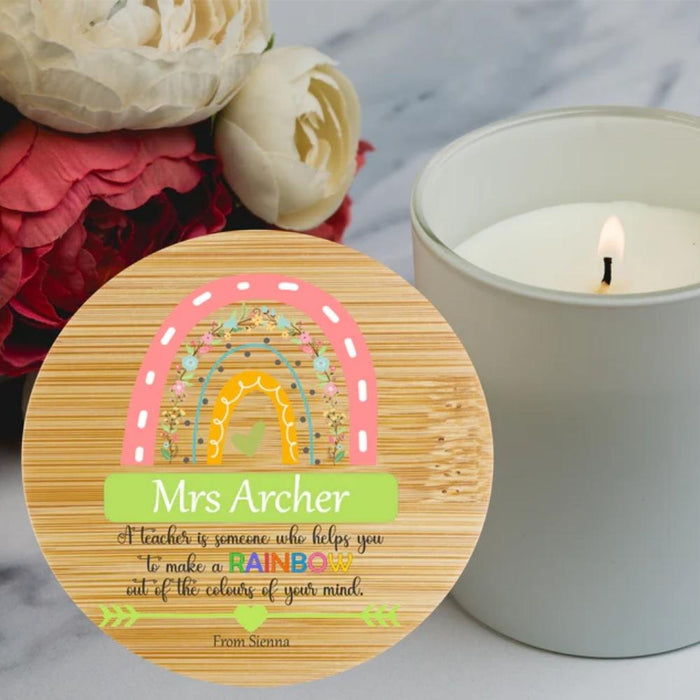 A round wooden bamboo candle keepsake with a colorful rainbow illustration and the personalised  text 'Mrs Archer A teacher is someone who helps you to make a RAINBOW out of the colours of your mind.' It is styled on a marble surface next to a lit candle and red flowers.