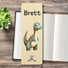 A personalized wooden bookmark featuring a cartoon green dinosaur with orange spikes, and a small volcano illustration at the bottom, displayed on a rustic wooden table next to an open book and a green succulent.