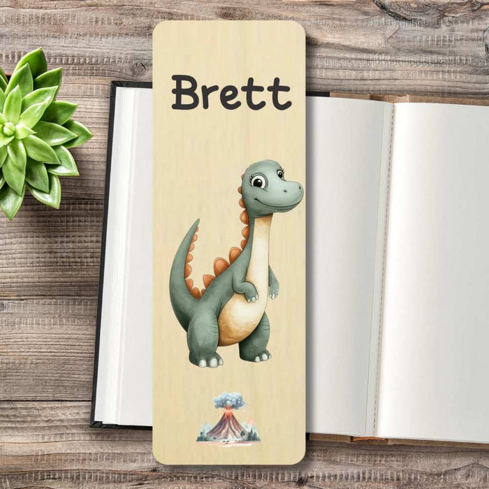 A personalized wooden bookmark featuring a cartoon green dinosaur with orange spikes, and a small volcano illustration at the bottom, displayed on a rustic wooden table next to an open book and a green succulent.