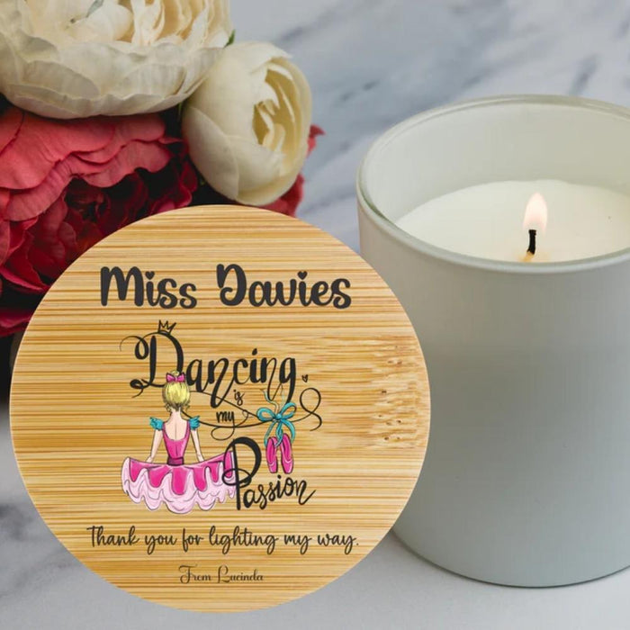 A round wooden coaster with a printed personalized design reading 'Miss Dawies Dancing my Passion' and 'thank you for lighting my way. From Lavender,' shown next to a lit white candle on a marble surface with flowers in the background.