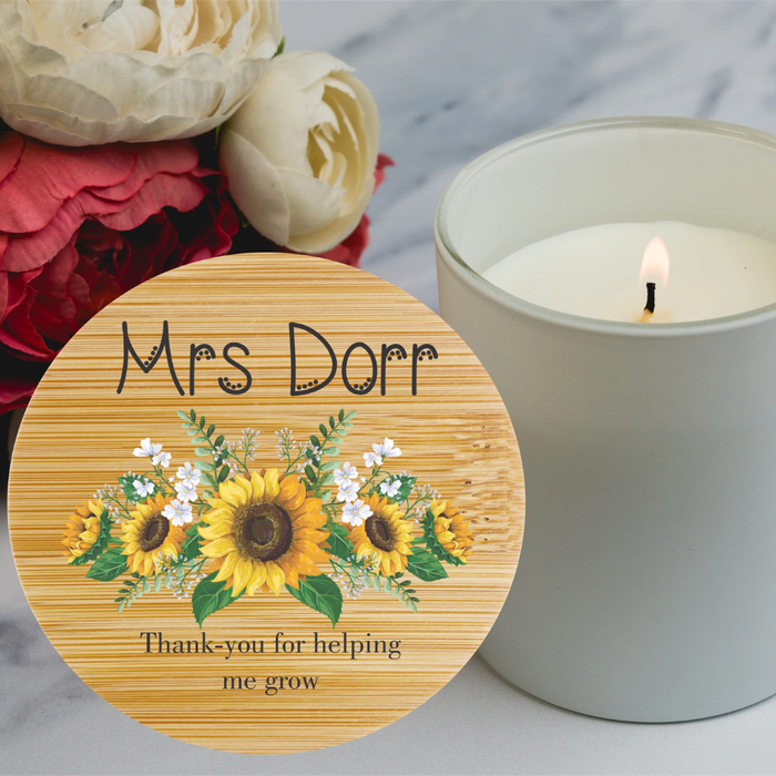 A round  lid for a personalised candle bamboo texture,  and a sunflower floral design, with the text 'Thank-you for helping me grow'. A lit white candle in a glass jar and a bouquet of red and white flowers are in the background on a light marble surface.