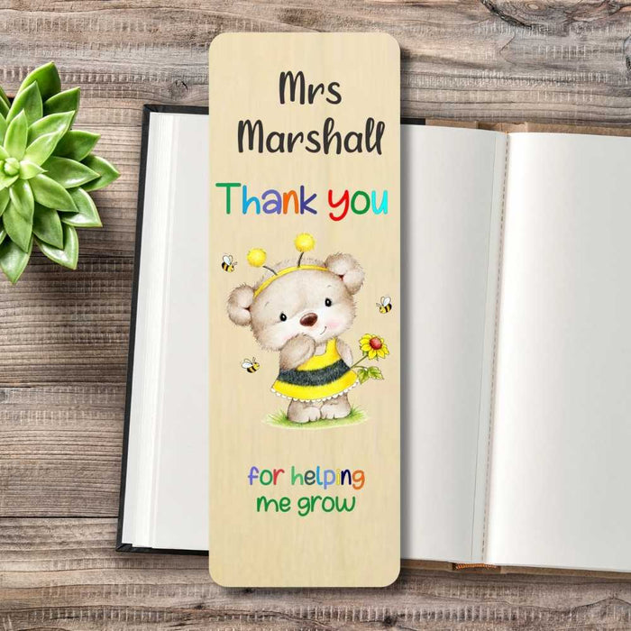 A personalized wooden bookmark featuring a cartoon bear dressed as a bee and the text 'Mrs Marshall Thank you for helping me grow', displayed on an open book with a small green succulent in the corner.