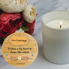 A round wooden keepsake tag with floral illustrations and the text ' It takes a big heart to shape little minds' is shown in the foreground next to a lit white candle on a light marble surface with red and white flowers in the background.