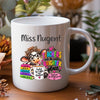 A white ceramic coffee mug with a colorful cartoon illustration of a girl with glasses and books, alongside the text 'Miss Nugent' and 'Teachers make the world a brighter place'. The mug is shown on a wooden table with a blurred background of plants and dried flowers.
