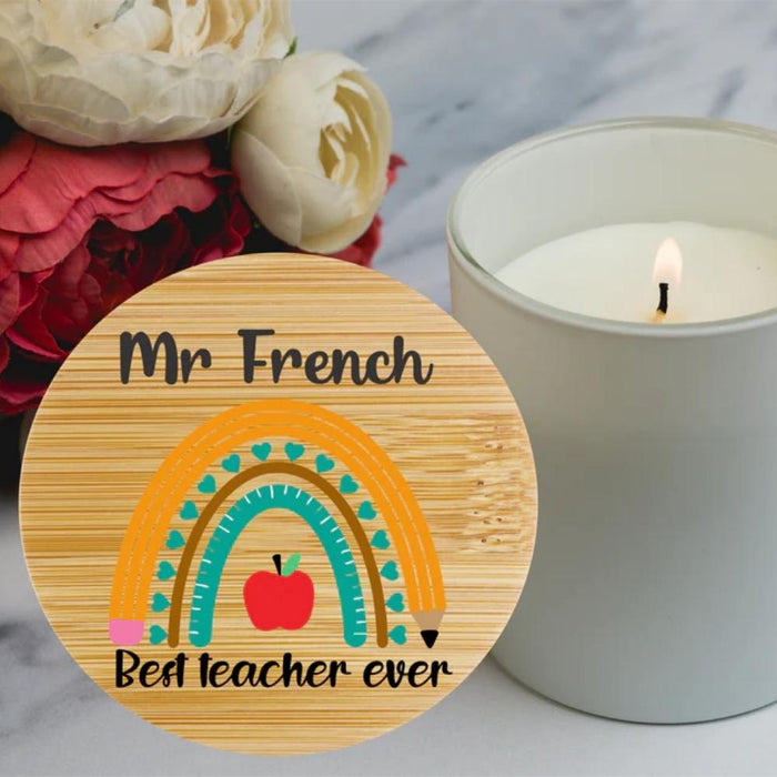A round wooden plaque or coaster with a printed rainbow, apple, pencil, and the text 'Best teacher ever', styled on a marble surface next to a lit candle and flowers.